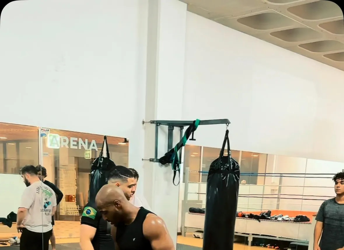 Sala de treino com sacos de boxe e ringue, com alunos a aquecer antes da aula